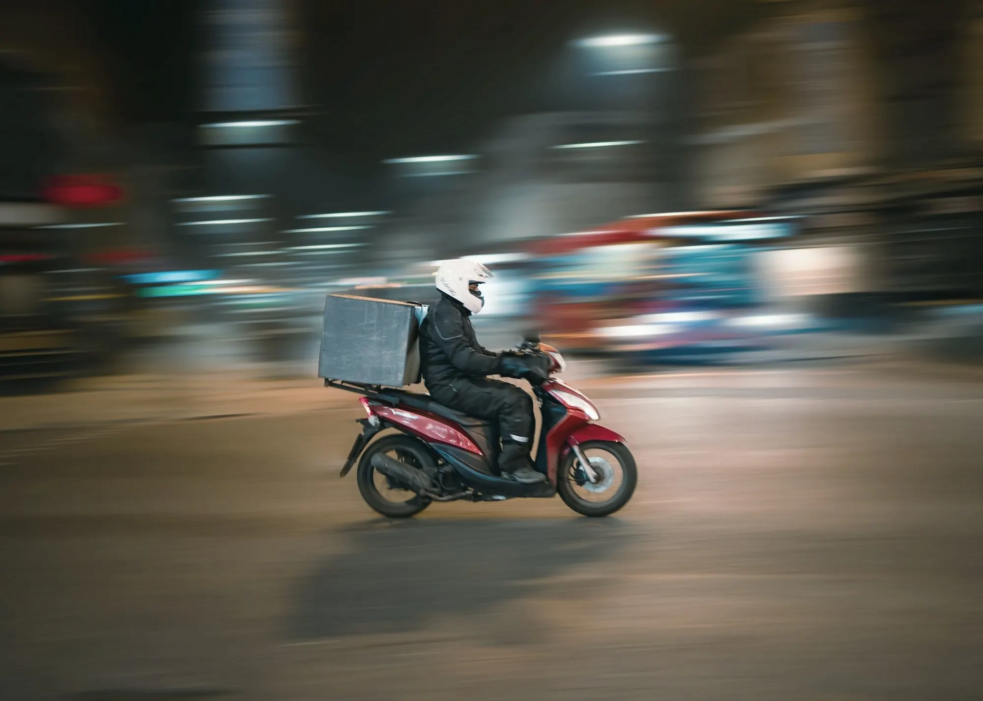 A delivery rider in a white helmet on a red scooter speeds past city lights.