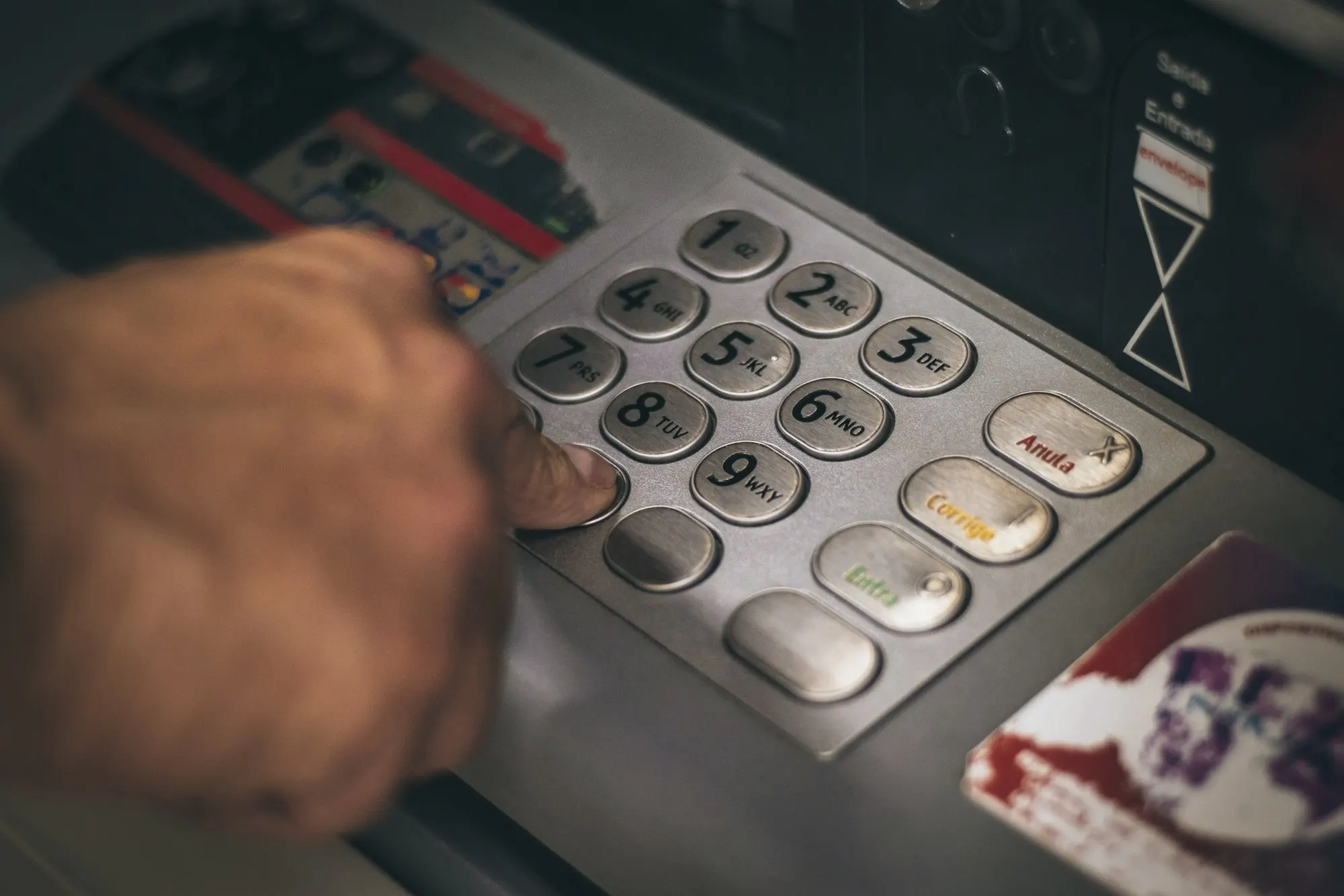 A finger presses a button on a metal ATM PIN keypad with numbered keys.